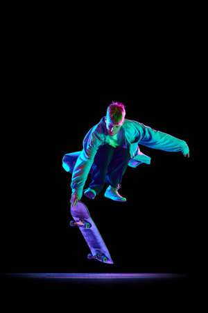 Dynamic image of young man in casual clothes in motion, training, skateboarding against black studio background in neon lightの写真素材