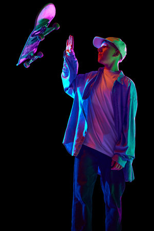 Boy in casual clothes standing with skateboard, posing, doing tricks against black studio background...