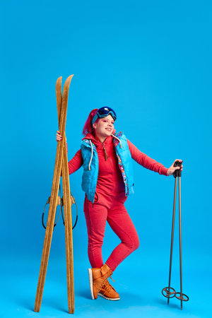 Young girl in colorful winter clothes standing with skis against blue studio background. Active holidaysの写真素材