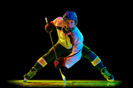Concentrated young woman, hockey player un uniform and helmet, standing with stick against black studio background in neon lightの写真素材