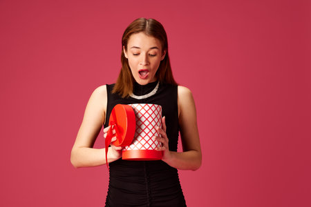 Surprise. Young beautiful woman in black dress, celebrating, opening present against pink studio...