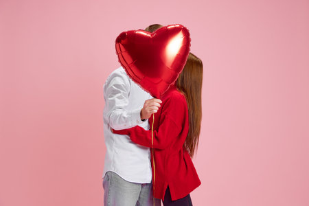 Happy young couple, woman and man holding balloon in heart shape and kissing behind against pink studio background. Valentines day, relationshipの写真素材