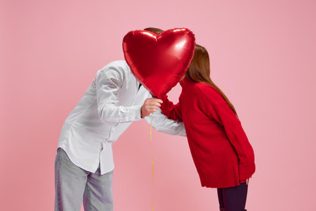 Happy young couple, woman and man holding balloon in heart shape and kissing behind against pink studio background. Valentines day, relationshipの写真素材