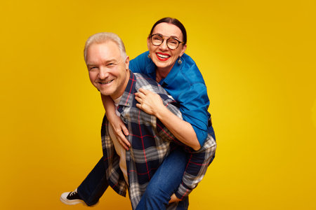 Beautiful, stylish, happy senior couple, man and woman having fun against yellow studio background. Woman on mans back. Valentines Dayの写真素材