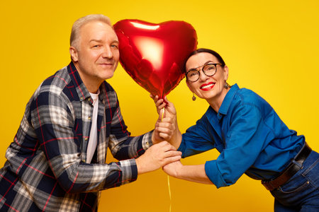 Beautiful, happy, senior couple, man and woman celebrating romantic holiday, holding balloon in heart shape against yellow background. Valentines Dayの写真素材