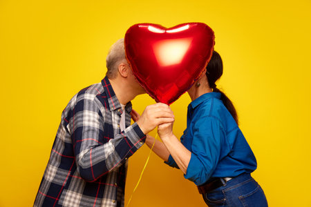 Senior couple, wife and husband standing behind red balloon in heart shape and kissing against yellow studio background. Valentines Dayの写真素材