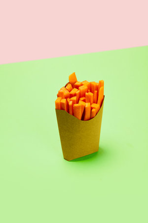 Cut, fresh carrots placed into carton packaging for fries against green background. Organic products and healthy foodの写真素材
