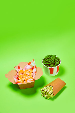 Healthy snacks. Green beans into popcorn basket, mandarin and celery against green background. Organic products and healthy foodの写真素材
