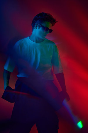 Handsome stylish young guy in white t-shirt and sunglasses posing over red background in neon...