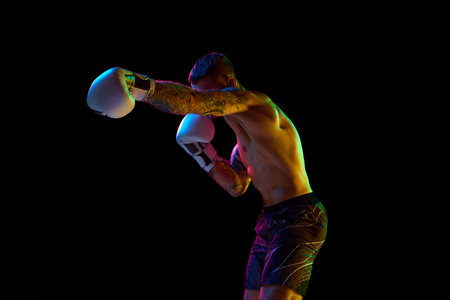 Young muscular man, boxing athlete in motion, training, fighting on ring isolated over black background in neon lightの写真素材