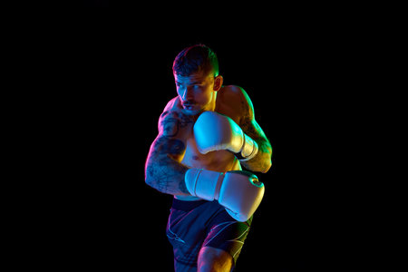 Competitive, strong, muscular young man, boxing athlete training shirtless isolated over black background in neon lightの写真素材