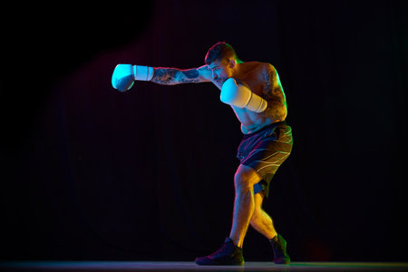 Strong, muscular young man, boxing athlete in gloves, training, fighting isolated over black background in neon lightの写真素材