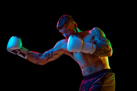 Strong, muscular young man, boxing athlete in gloves, training, fighting isolated over black background in neon lightの写真素材