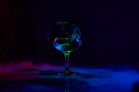 Martini splash. Glass with popular cocktail standing against dark background with neon light with smokeの写真素材