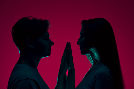 Silhouette of young man and woman standing closely to each other, holding hands, expressing love against purple background in neon lightの写真素材