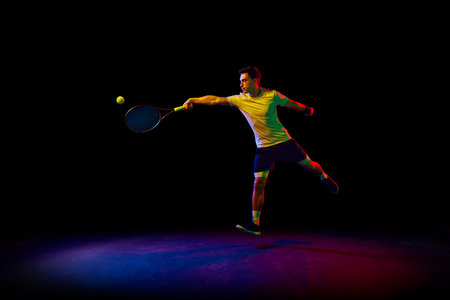 Concentrated and competitive man, tennis athlete during training session, hitting ball with racket against dark background in neon lightの写真素材