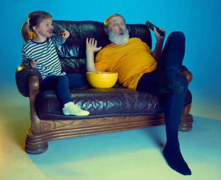 Cheerful little girl watching TV at home with her loving grandfather over blue background in neon lightの写真素材