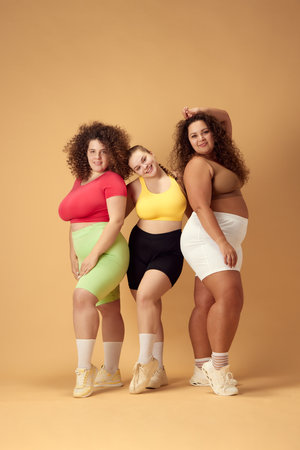 Portrait of three beautiful young women with overweight body standing in sportswear, smiling over beige studio backgroundの写真素材