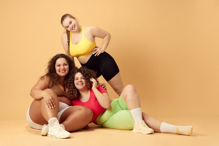 Three beautiful young women, plus size models with oversized bodies, posing in sportswear over beige studio backgroundの写真素材