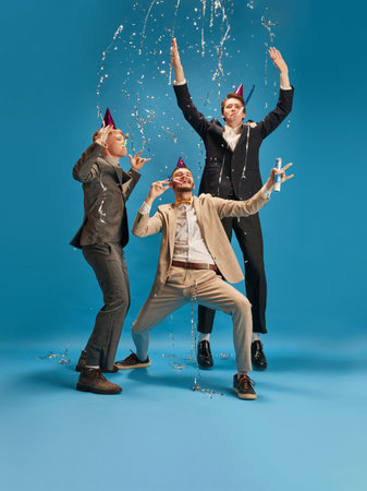Emotional young people, men in elegant suit celebrating, having fun against blue background. Merry...