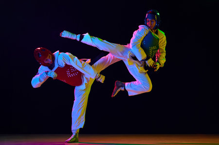 Dynamic image of young men, taekwondo athletes in kimono and helmets training against black...
