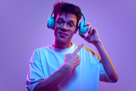 Young guy in glasses using headphones, pointing on it with finger against purple background in neon light. Good sound, high quality accessoryの写真素材