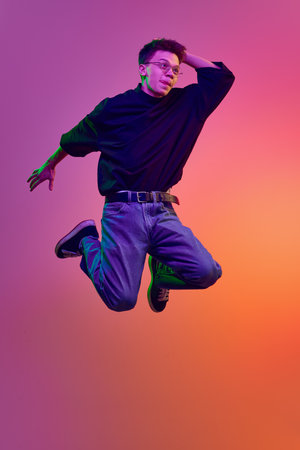 Young man in casual stylish clothes and eyeglasses jumping with positive face against gradient pink background in neon lightの写真素材