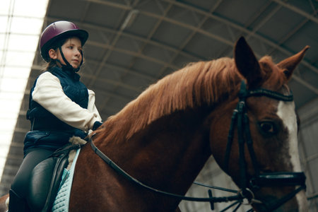 Portrait of little girl, child in helmet sitting on brown horse, training, practicing horseback riding.の写真素材