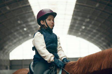 Portrait of little girl, child in helmet sitting on brown horse, training, practicing horseback riding.の写真素材