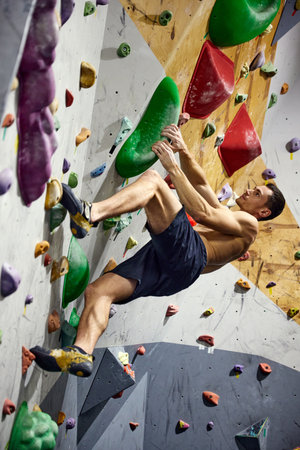 Shirtless, muscular young man climbing artificial rocks on climbing wall, practicing bouldering activity indoors.の写真素材