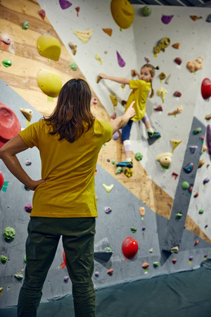 Female bouldering trainer teaching little girl, child bouldering activity, techniques. Little girl climbing wall, indoor class