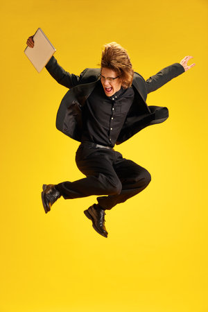 Young man, dressed smart casual, fashion and glasses outfit jumping of joy and raising hands holding laptop against yellow background.の写真素材