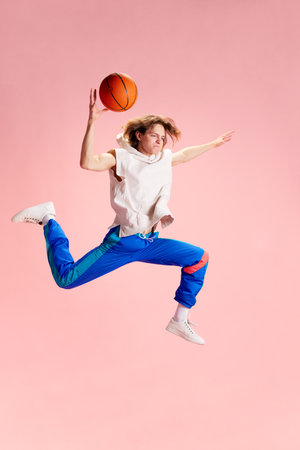 Young man dressed in retro sportwear sitting and posing with basketball ball against pastel pink...