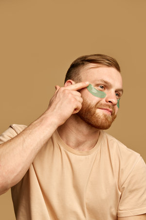 Close up portrait of young attractive man applies hydrogel patches under the eyes against swelling and bruising against beige background.の写真素材