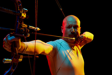 Portrait of serious man, archer aiming with bow on target against black studio background in neon...