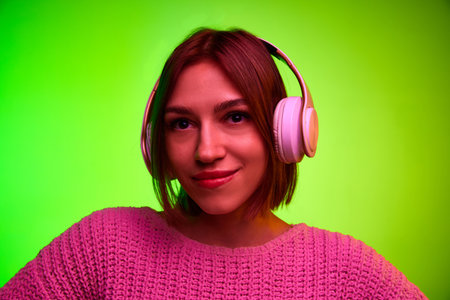 Close up portrait of young girl with headphones on head looking at camera against gradient green...