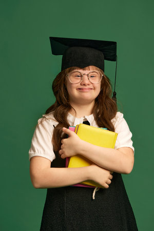 Inclusive education campaign. Girl with down syndrome standing with books, wearing graduation cap against green studio backgroundの写真素材