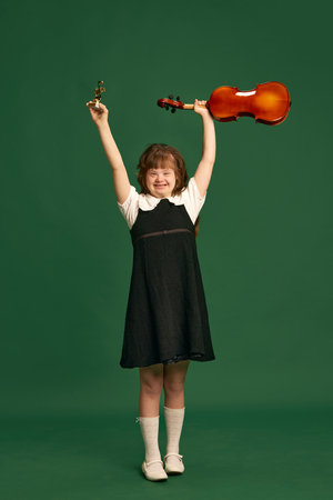Beautiful girl with down syndrome standing with violin, learning music against green studio backgroundの写真素材