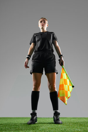 Young woman, female referee in uniform standing with flag, controlling game rules against grey...