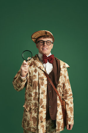 Young man wearing vintage clothes, standing with magnifying glass against green studio background. Detective storyの写真素材