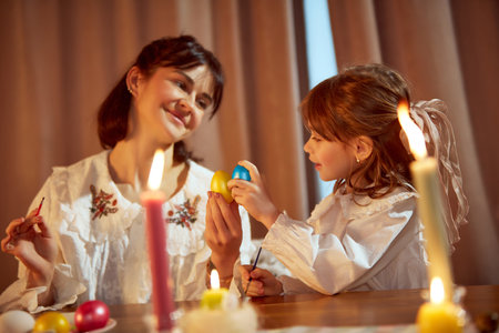 Magic cozy moment. Beautiful mother and her little daughter paining eggs, preparing for Easter...