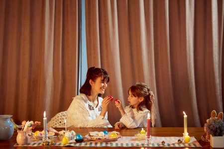 Cracking eggs for a good luck. Smiling happy mother and her little daughter holding painted Easter eggs. Home celebration and Easter traditionsの写真素材