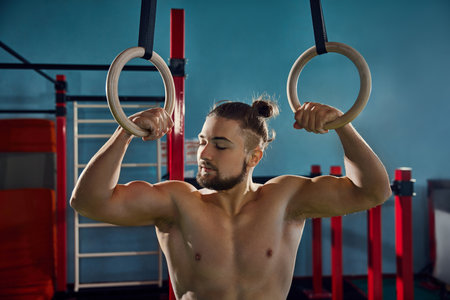 Shirtless, athletic young man showing his muscular, strong body, training with gymnastic rings in...