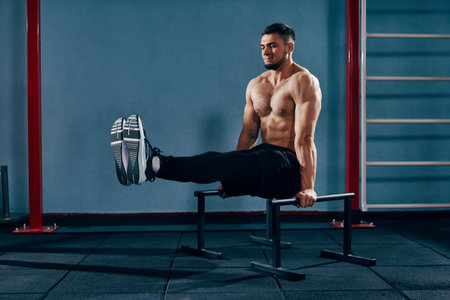 Full-body workout. Muscular, shirtless athletic man training in modern gym, doing L-sit exercise on...