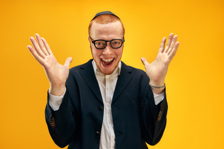 Portrait of happy young redhead Jewish man in glasses and yarmulke cheerfully posing against yellow studio backgroundの写真素材
