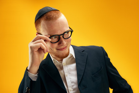Portrait of young redhead Jewish man in yarmulke, glasses and suit smiling, posing against yellow studio backgroundの写真素材