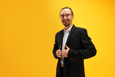 Portrait of emotional, excited Jewish man in black suit, yarmulke posing against yellow studio background. Happy holidaysの写真素材