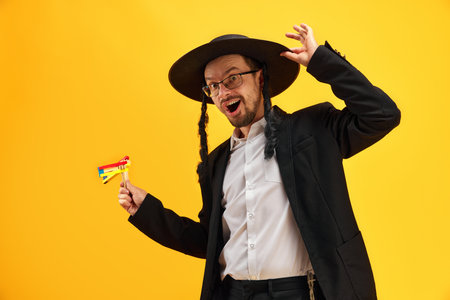 Cheerful Jewish man in hat, with sidelocks holding noisemaker, against yellow background. Celebrationの写真素材