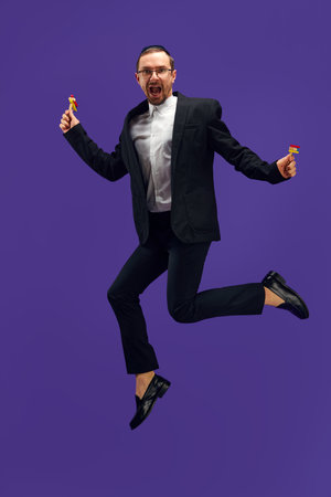 Full-length of emotional Jewish man in his 30s wearing black suit, holding noisemaker and jumping against purple studio backgroundの写真素材