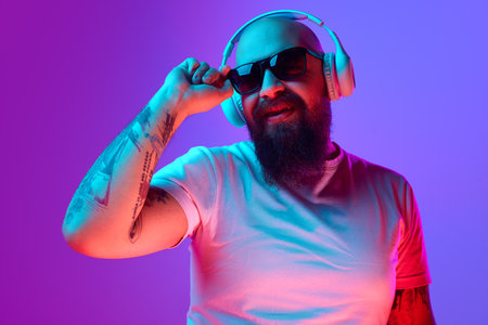 DJ. Bearded, artistic bald man with tattoos, in white t-shirt and sunglasses standing against gradient purple background in neon lightの写真素材